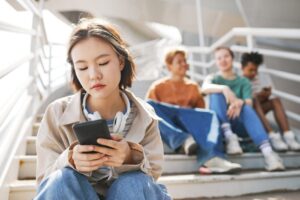 Young woman sits on outdoor stairs looking at her smartphone, headphones around her neck, while a group of friends sits blurred in the background.