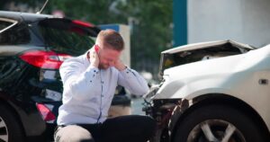 Man in a light shirt sitting beside a white damaged car, hands on head in distress after a crash in a street scene