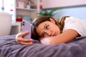 Young woman lying on a bed, looking at her lavender smartphone in her hand.