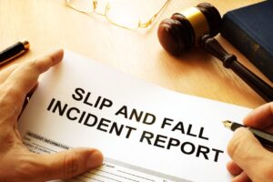 Close-up of a hand holding a slip-and-fall incident report form on a desk, with a gavel and glasses nearby indicating legal/administrative review.