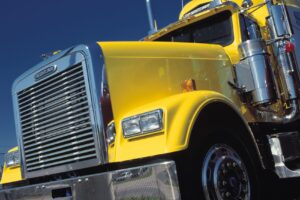 Close-up of a bright yellow Freightliner truck cab from a low-angle side view, showing chrome grille and headlight details against a blue sky.