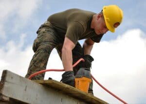 Construction worker in a yellow hard hat using a power tool on a wooden beam outdoors.