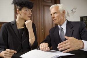Two business professionals, a woman and an older man, discuss documents at a desk in an office setting.