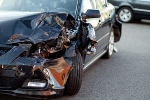 Front-end damage to a dark car after a collision, with crumpled hood and broken headlight visible on the left side in a parking lot or street setting.
